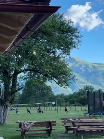 A group of people are playing volleyball in a grassy field. There is a large tree to the left of the frame, and mountains in the background. A wooden picnic table with benches is in the foreground. The sky is blue with some clouds. A trampoline is visible on the right side of the frame.