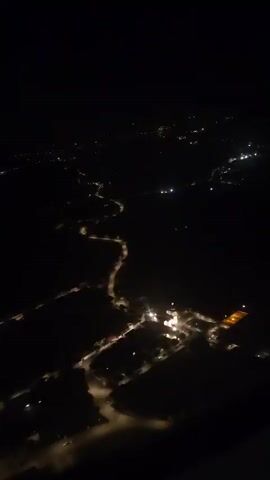 The video is a night-time aerial view of a winding road with scattered lights from buildings and streetlamps. The landscape is dark, with the lights creating illuminated patches and outlining the curves of the road. The view is from a high vantage point, possibly an airplane, looking down on a rural or semi-rural area. The lights suggest human settlement along the road.