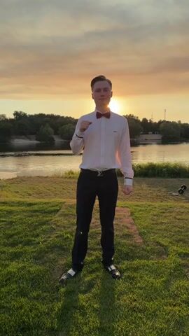 This media file features a young man standing in a grassy field by a river at sunset. He is wearing a white dress shirt, black pants with a distinctive belt buckle, a red bow tie, and black and white shoes. The sun is low in the sky, casting a warm glow and creating a silhouette effect on the trees in the background. The sky is filled with soft, orange and yellow hues, indicating a pleasant evening. The river is calm, reflecting the colors of the sunset. The man appears to be posing or performing a dance, as his hands are in motion. He has a neutral expression. There is some faint text on his shirt that appears to be a logo. The setting suggests an outdoor event or photoshoot in a natural landscape.