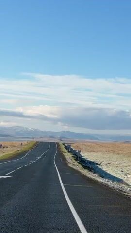 The video shows a road stretching into the distance under a clear blue sky with scattered clouds. In the background, mountains are visible. The road is black with white lane markings. On either side of the road is a field of dry grass with some frost. There are several road signs along the right side of the road. The video is taken from a moving vehicle.