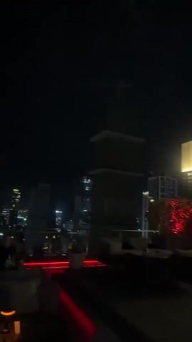 The video depicts a nighttime view of a city from a rooftop setting. The scene is characterized by the glow of city lights against a dark sky, suggesting it is after sunset. In the foreground, there are people seated at tables, with some visible objects that appear to be tables and seating arrangements. Red accent lighting is present, adding to the ambiance of the location.

The background showcases a skyline filled with numerous buildings of varying heights, many of which are illuminated, indicating an urban environment. The lights create a blurred, bokeh effect, common in nighttime photography and videography. The overall impression is that of a lively urban area at night, possibly a restaurant, bar, or entertainment venue with a view.

The location context provided is Khan Boeng Keng Kang, Cambodia. This suggests the cityscape being viewed is that of Phnom Penh. The time of day is clearly night. There are no strong indicators of weather, but the darkness suggests clear skies. The emotions evoked are likely those associated with a night out, possibly relaxation, enjoyment, or socializing, given the presence of people in a seemingly social setting. There is no visible text within the video frame.