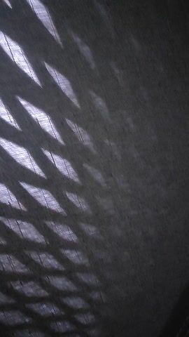 This media file depicts an indoor scene with a focus on a textured surface, possibly fabric or leather, with diamond-shaped light patterns cast upon it. The lighting appears to be artificial and casts shadows that create a geometric, almost gridded effect. In the upper right portion of the frame, a piece of lined paper is visible with handwritten text in blue ink. The text is partially obscured and angled, but legible words include "ME," "bet: over," "2.0," and "Coef: 1.68." The overall impression is one of a close-up view of an object or surface, with an interesting interplay of light and shadow. The presence of the paper with writing suggests it might be a note or some form of information being displayed or referenced in the scene. The color palette is dominated by dark blues and grays, with the white of the paper providing a contrast. There are no people visible in the frame. The location context of Xuân Long, Vietnam, does not provide any direct visual cues within the image itself.