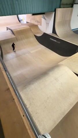 A person wearing a helmet and all black clothing is riding a scooter on a plywood skatepark. The camera follows them as they ride down a small ramp, up another ramp, and then down into a larger ramp. They turn and ride up a large wall. The skatepark has the words "Adrenaline Alley" and "Fossil Skate" on the wall.