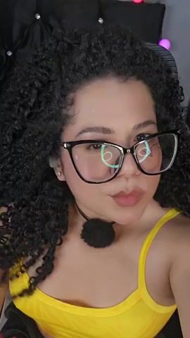 A close-up shot focuses on a young woman with curly dark hair, wearing large black-rimmed glasses and a yellow tank top. She has a fluffy black pom-pom choker around her neck. The background is dark and out of focus, with some indistinct colorful lights visible. The woman pouts her lips and looks directly at the camera. She has a small tattoo on her left arm and her right hand is visible near the bottom of the frame. The lighting is soft and diffused.