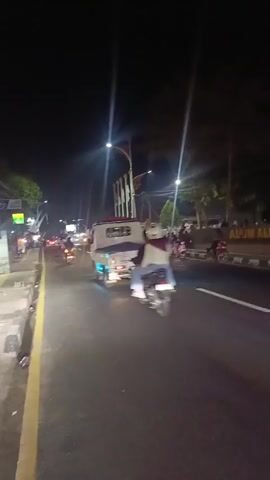A nighttime scene on a road shows several motorbikes and a three-wheeled vehicle. The vehicles are moving, and some have their brake lights illuminated. On the side of the road, there is a sign that appears to say "ALUN ALUN KALIJATI" and several flags. Trees and streetlights are visible in the background. Some people are on the motorbikes, and others appear to be standing on the side of the road. The lighting is dim, with bright streetlights creating lens flares.