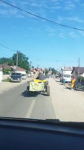 The video shows a view from inside a vehicle looking forward on a paved road. In the foreground, the dashboard of the vehicle is visible. The road is lined with houses and trees on both sides. In the center of the road, a person is riding on a small, yellow trailer attached to a tractor or similar vehicle. The trailer is covered with a green tarp. Several other vehicles, including a white van and a dark-colored van, are parked on the side of the road or driving in the distance. The sky is clear and blue, indicating a sunny day. The scene appears to be in a rural or suburban area.