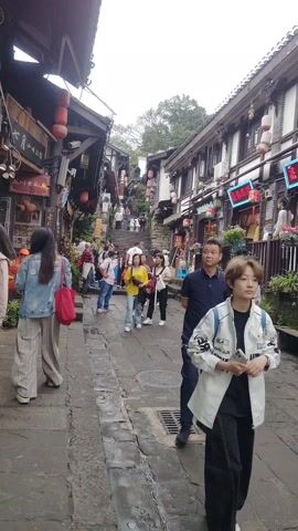 The video shows a crowded street in a traditional Chinese town. The street is paved with grey stone. Buildings line both sides of the street, with shops displaying various goods. Red lanterns hang from the eaves of the buildings. People are walking up and down the street. Some are looking at the shops, while others are just passing through. There is a set of stairs at the end of the street, leading uphill. The overall atmosphere is busy and touristy.