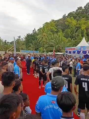 This media file captures a lively outdoor event, likely a sporting ceremony or tournament, set in a tropical location. A crowd of people, dressed in a mix of casual wear and team uniforms, fills the scene. Many are wearing bright blue t-shirts with the text "KAPOLRES CUP 2023" visible, suggesting a connection to a police-sponsored cup. Some individuals are in black and white football kits, indicating they might be participants or team members.

The event appears to be taking place on a grassy field, with a prominent red carpet laid out, leading towards a stage or a designated area. Several white tents are erected in the background, one of which displays banners with text. Visible text on these banners includes "KAPOLRES CUP PI DIE JAYA" and "8 THUN 2026," and "HARI BAYANGKARA." These suggest the event is related to the "Hari Bayangkara" (Police Day) celebration and possibly a football tournament called the "Kapolres Cup" in the "Pidie Jaya" region, with the year 2026 mentioned on one banner.

The atmosphere seems festive and energetic, with people interacting and seemingly engaging in a pre-event gathering or a celebratory procession. The presence of individuals in police uniforms suggests official involvement. The background features lush green trees and hills, characteristic of a tropical setting, and the lighting indicates it is daytime, likely late afternoon, given the slightly diffused sunlight. The overall scene conveys a sense of community participation and local celebration.