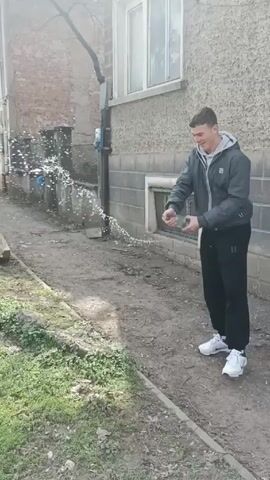 A young man is standing in a narrow alleyway between two buildings. He is wearing a gray hooded jacket, black pants, and white sneakers. He is holding a white object in his hands, and he is shaking it vigorously. This causes a spray of white liquid to erupt from the object and scatter around him. The alleyway is cluttered with dirt, grass, and debris. The buildings are old and made of brick and concrete. The weather appears to be overcast.