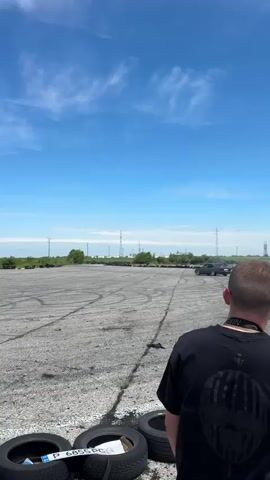 The video shows a gray BMW E34 drifting on a paved area marked with tire tracks. The car enters the frame from the right, spins around, and exits left, creating a cloud of smoke. In the foreground are several tires. A person in a black shirt is visible in the lower right corner, apparently filming the action. The background includes power lines and stacks of tires, with a clear blue sky overhead dotted with some clouds.