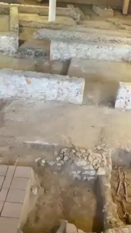 The video shows an archaeological excavation site. The site appears to be indoors, possibly a museum or a protected area. The camera pans across the site, revealing several excavated areas. One excavation reveals a human skeleton, lying in what appears to be a grave. Surrounding the grave are various stones and brick structures, presumably the remains of an ancient building. In the background, a section of mosaic floor is visible. Metal scaffolding is also visible, providing structural support to the site.