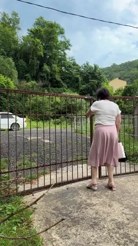 A woman with dark hair, wearing a white t-shirt and a pink skirt, opens a rusty metal gate. She is seen from behind, as she pushes the gate open with her right hand and reaches for a bag with her left. Her left leg is slightly bent. She is wearing sandals with light-colored heels. The gate is made of vertical metal bars and appears old. Beyond the gate, there is a gravel area and lush green foliage, suggesting an outdoor, possibly rural setting. Trees and a hillside covered in greenery form the background. A white car is visible to the left, parked behind the gate. A building with a yellow facade can be seen on the hillside in the distance. The sky is overcast, indicating possibly cloudy or rainy weather. The scene seems to be taking place during the daytime. The woman's action suggests she is entering or exiting a property. There is no visible text in the media.