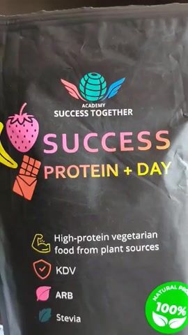 The video shows a close-up of a bag of "SUCCESS PROTEIN + DAY" powder. The bag is black with colorful graphics and text. Visible on the bag are images of a strawberry, a banana, and a chocolate bar. The text on the bag includes "ACADEMY SUCCESS TOGETHER", "SUCCESS PROTEIN + DAY", "High-protein vegetarian food from plant sources", "KDV", "ARB", "Stevia", "sugar free", "gluten free", "VEGAN", and "30 sticks". There is also a green circular sticker that says "NATURAL PRODUCT 100%". The bottom of the bag has a pink and orange band with text that appears to list flavors, including "YUMMY CHOCOLATE" and "JUICY STRAWBERRIES". The lighting suggests the bag is on a table or shelf, possibly in a home or retail environment.