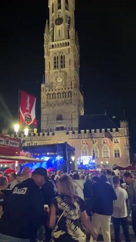 A large crowd of people are gathered in a square at night, facing a tall, brick tower. There is a stage set up in front of the tower with a large screen displaying a live feed of a concert or event. Flags are visible on the tower and surrounding buildings. People are dressed in casual clothing and appear to be enjoying the event. The atmosphere is festive, with lights and music. The screen on the right side of the frame shows a bright, colorful image of a performance with many people in attendance, possibly a concert. The screen on the left side of the frame shows abstract blue and white geometric shapes. The tower is illuminated, and there are streetlights in the square. The overall scene depicts an outdoor gathering or festival.