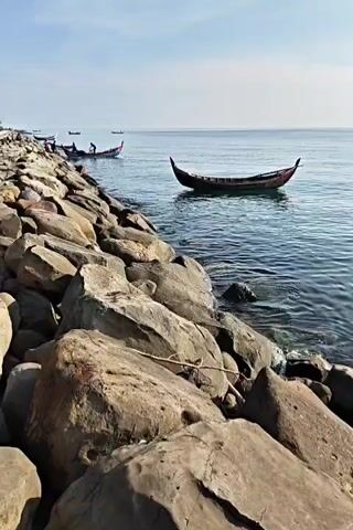 The video shows a rocky shoreline on a sunny day. Several small boats are visible in the water. Some appear to be rowboats with people on board. One boat is closer to the camera and appears to be floating on the water near the rocks. The water is relatively calm. The sky is mostly blue with some wispy clouds.
