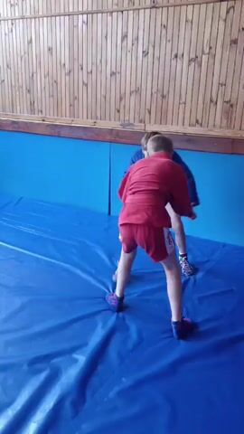 Two young boys, dressed in wrestling attire, are engaged in a Sambo training session on a blue mat. One boy wears a red Sambo uniform, and the other wears a blue Sambo uniform. The setting appears to be an indoor training facility with wooden paneled walls in the background. The mats are spread out, indicating a space dedicated to combat sports. The boys are in mid-action, demonstrating throws or grappling techniques. The lighting suggests it's daytime, possibly indoors with artificial lighting supplementing natural light. The overall atmosphere is one of athletic training and skill development. No visible text is present in the frame.
