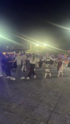 The video shows a night scene with a group of people holding hands and dancing in a circle. Several individuals are wearing traditional folk costumes with colorful patterns and white blouses or shirts. They are on a paved outdoor area, possibly a town square, with buildings and trees in the background. Bright lights illuminate the scene, creating lens flares. Some people in the background appear to be spectators. The overall atmosphere seems festive and celebratory.