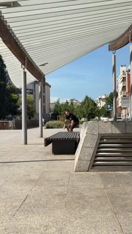 A skateboarder wearing a black t-shirt and brown pants performs a trick. The skateboarder approaches a dark wooden bench, jumps onto it, and then jumps off the far side. The skateboarder's left arm is extended, and their right arm is bent. The skateboard is yellow with a graphic design. The scene takes place outdoors on a sunny day. The background includes trees, buildings, and a covered walkway with white slats. The ground is paved with gray stones. The skateboarder lands on the ground and the skateboard is visible in mid-air.