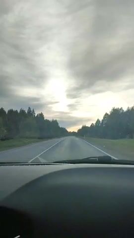 The video shows a car's point of view while driving on a straight asphalt road flanked by dense green trees on both sides. The sky is overcast with a pale sunlight shining through, creating a bright spot in the center of the view. The road is marked with a white line in the middle. The car's dashboard is visible in the foreground. Some water streaks can be seen on the windshield, suggesting recent rain. In the background, the road appears to slope slightly upwards. The car appears to be moving steadily on the road.