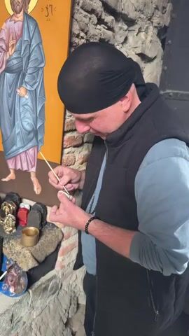 A man wearing a black head covering and a dark vest is standing and using a thin wooden stick to handle a small piece of white material. Behind him is a large religious painting of a man in a blue robe. Below the painting is a stone alcove with various small religious icons, paint tubes, a brass cup, and a painter's palette. The background wall is made of rough stone and brick.