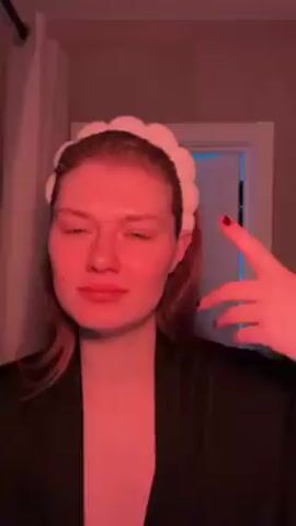 A young woman stands in a bathroom with a pinkish red cast from the lighting. She's wearing a black satin robe and has a white, textured headband holding back her hair. Behind her is a doorframe with a white door, and an open doorway reveals a bright blue or white light coming from inside. She leans her head back and stretches her neck. A white curtain is visible to her left.