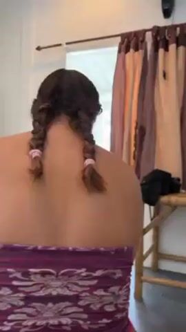 A woman with brown hair styled in two braids is standing in a room. She's wearing a strapless purple top. The room has a door, bamboo furniture, and curtains. She turns her head, smiles, and looks at her phone.
