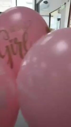 Here are the details about the video. The video shows a close-up shot of three pink balloons. The central balloon has "It's a Girl" printed on it in gold lettering with a heart design. To the left of the central balloon is a pink balloon with white heart designs. To the right of the central balloon, another pink balloon is visible with the text "Добре дошло" and small white stars on it. The background includes a blurred view of a window, and parts of a building are seen outside the window. Additionally, there is what appears to be a menu or board partially visible behind the balloons.