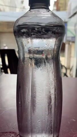 A close-up shot focuses on a clear plastic bottle filled with water, its surface covered in condensation. The bottle is placed on a reddish-brown surface, possibly a table. In the background, blurred elements suggest an outdoor or semi-outdoor setting, with hints of buildings and possibly vehicles. The water inside the bottle appears to be agitated, creating a swirling effect.