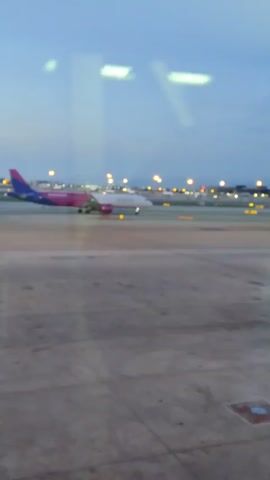 From the perspective of someone inside, the video shows a view of a plane on a runway at an airport. It is dusk or dawn, the sky is a light blue, and the runway is visible below, with lights illuminating the airfield. The plane is painted white, purple and blue. The recording then moves, ending on an obstructed view of a gray wall.