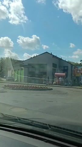 This video shows a roundabout in what appears to be a European town on a sunny day. In the foreground, we see the dashboard and windshield of a car, indicating the perspective of the viewer. In the middle ground, a circular flower bed is visible, surrounded by a paved road. Beyond the roundabout, a building with green signage can be seen, possibly a shop or cafe. Several people are gathered in front of this building, near a red awning that appears to be covering a market stall. There are also cars and a street sign visible in the background. The sky is blue with scattered white clouds, suggesting fair weather. The overall scene depicts a typical street view in a small town.