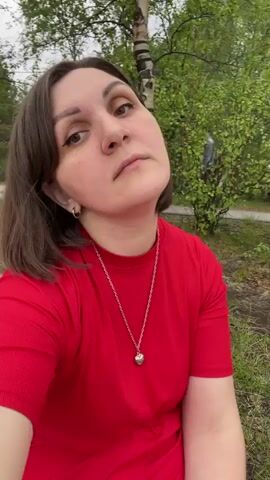 A woman wearing a red shirt and a silver necklace sits on grass in an outdoor setting. Her head is slightly tilted as she looks at the camera, and her facial expression is neutral. In the background, there are trees and a red structure that appears to be a gazebo or shelter. There are also some picnic blankets on the grass to her right. Another person is visible in the background, standing near the trees. The lighting suggests it is daytime.