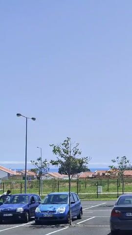 The video captures a bright, sunny daytime scene in a parking lot in Ponta Delgada, Portugal. A clear, light blue sky dominates the upper portion of the frame.

In the foreground, two blue cars are parked. On the left, a dark blue Renault Clio hatchback (license plate: 23-75-IJ) is shown, with a person getting into the driver's seat at the beginning of the clip. To its right, a light blue Renault Clio hatchback (license plate: 81-69-UP) is parked.

As the video progresses, a white Volkswagen Golf hatchback (license plate: AU-19-MG) drives from the right side of the frame towards the left, passing in front of the parked blue cars. The person entering the dark blue Clio closes the door as the white Golf drives by.

In the mid-ground and background, there are several streetlights, small green trees, and a fence. Beyond these, residential buildings with orange and yellow tiled roofs are visible, characteristic of the region. On the distant horizon, a faint blue line indicates the presence of the ocean under the clear sky. The overall scene depicts a mundane moment in a sunny, coastal setting.