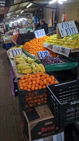 The video shows a produce stand in a market. There are several tables laden with various fruits, including bananas, oranges, avocados, tomatoes, kiwis, lemons, limes, and grapes. Each table has a sign displaying the price of the fruit, most marked at $1000 or $2500. The fruit is arranged in neat piles and rows. A few people are visible in the background, seemingly shopping or working at other stalls. The market has a high ceiling with fluorescent lighting and exposed beams. The floor is a worn wooden surface. Black plastic crates are stacked underneath the tables.