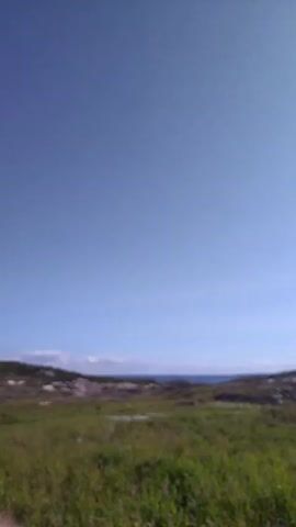 This is a video of a landscape with a clear blue sky above. In the foreground, there is green grass with some scattered rocks. In the middle ground, there is water and rocky hills. In the distance, there appear to be buildings and more hills. The sun is shining brightly in the upper right portion of the frame, creating some lens flare. There are a few scattered clouds in the sky. The video pans slightly to the right, revealing more of the coastline and the sun. There are no people or animals visible in the video. There are some man-made objects visible, including what appears to be a wooden structure in the lower right corner and possibly a small building on the distant shore.