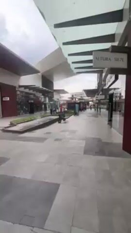 The camera is positioned at ground level in a modern outdoor shopping mall on an overcast day. The camera moves forward along the walkway. The mall features a mix of architectural styles, including flat roofs, angled ceilings, and dark red accents. Several benches are placed along the walkway, and planters with greenery line the sides. Storefronts with large glass windows are visible on either side, reflecting the environment. A sign reading "ALTURA SIETE" is visible on the right side of the frame. Several people can be seen walking in the distance, appearing as small figures within the mall's open space. The ground is paved with large grey tiles in a geometric pattern.