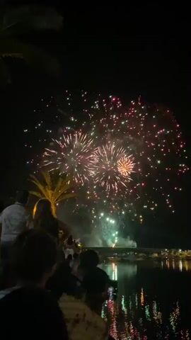 The video captures a lively nighttime scene in Tavira, Portugal, filled with the excitement of a fireworks display. In the foreground, silhouetted figures of people are gathered, their backs to the camera, suggesting they are watching the spectacle unfold. A palm tree is visible on the left, hinting at the warm climate.

The main event is a vibrant fireworks show illuminating the dark sky with bursts of color, including pink, red, and green. The reflections of the fireworks and distant lights shimmer on the calm water in the foreground, creating a mesmerizing visual effect. On the opposite side of the water, a waterfront promenade is lined with buildings and illuminated with lights, adding to the festive atmosphere.

The middle section of the video briefly shows a blurred shot of distant lights, likely from the town or the fireworks launch site. This is followed by close-ups of a woman smiling and looking up at the fireworks, her face lit by the explosions in the sky. Her joyful expression conveys a sense of wonder and enjoyment.

The overall mood is celebratory and engaging. The time of day is clearly night, and the weather appears clear, allowing for a spectacular view of the fireworks. The combination of the fireworks, the waterfront setting, and the people's reactions creates a memorable and festive occasion. No visible text is present in the video.