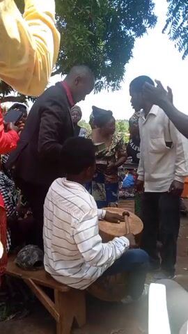 A group of people stand in a circle outdoors, seemingly in a celebration or event. In the center of the group, a young person sits on a wooden stool and plays a drum. Surrounding them are several people, some clapping and moving to the rhythm. A man in a suit appears to be leading or participating in the activity, holding what looks like a small book or phone. Other individuals are dressed in casual or traditional clothing, some holding phones to record. The setting includes a tree and glimpses of the surrounding landscape in the background.