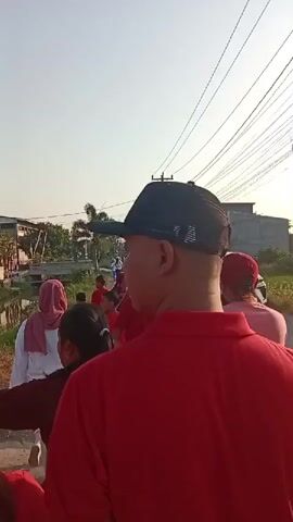 A group of people, many wearing red, are walking on a dirt road next to a canal or ditch. The sun is low in the sky, creating a bright glare. Some people are in the foreground, their backs to the camera, while others are further down the road. There are buildings and trees in the background, as well as power lines. A white car is parked on the side of the road. A motorcycle is also visible. The overall atmosphere seems to be one of a procession or a community gathering.