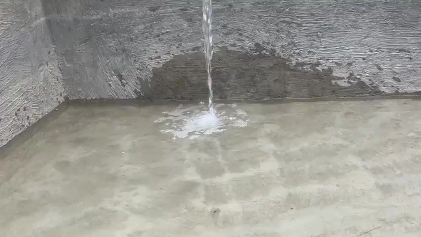 Water is flowing from a pipe and splashing into a concrete container, creating ripples and bubbles on the surface of the water. The container walls are made of rough, grey concrete. The video shows a close-up view of the water flow and then pans out to reveal more of the container.