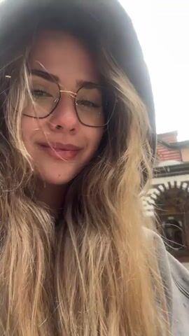 A young woman with long blonde hair wearing glasses and a gray hoodie stands in the courtyard of a monastery. She smiles at the camera and then turns to show the monastery buildings, which have a distinctive striped design with a dome. She then turns back to the camera with a surprised expression. Other people can be seen in the background. The weather appears to be overcast.
