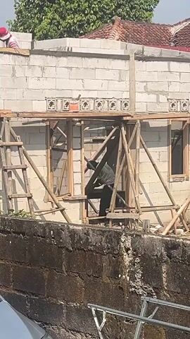 The video shows a construction site. In the foreground, a stone wall is visible. In the midground, a building under construction is seen. The building is made of concrete blocks and has scaffolding made of bamboo poles. A person wearing a grey shirt and black pants is working inside the building, near a window opening, using tools. Another person wearing a red hat and striped shirt is on top of the wall, observing. Trees and a red-tiled roof are visible in the background. The lighting suggests it is daytime.