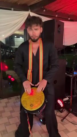 A young man with curly brown hair and a beard, wearing a black jumpsuit and a multicolored ribbon around his neck, plays a yellow hand drum. He is standing on a tiled floor in what appears to be an outdoor venue with white draping and straw-covered roofing. There are large speakers, stage lights, and other musical equipment in the background. The man is actively engaged in playing the drum, with his hands in motion and his head tilted back at one point, suggesting he is immersed in the music. The lighting is dim and colorful, indicating it is likely nighttime or indoors.