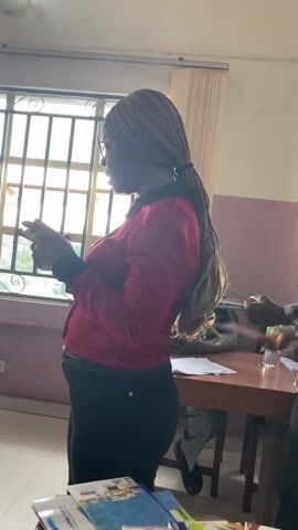 Here is a description of the video:

The video shows a woman wearing a red blouse and black pants standing in a room and talking, holding a stack of small boxes in her hands. She has braids and wears glasses. There is a table in front of her with more boxes and a bottle of water on it. A man in a plaid shirt is partially visible behind her to the right.  Another man is seated at a table in the background, writing on a paper. The room has a window with bars. The woman appears to be giving a presentation or explaining something about the items she is holding.