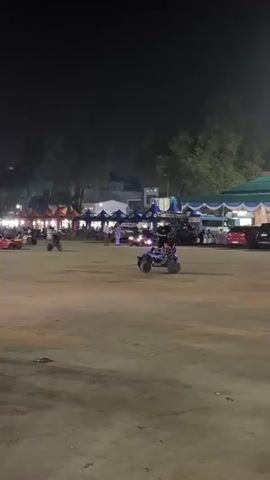 This is a vertical, nighttime video clip from an outdoor event, likely a fair or festival, in Lubuk Pakam, Indonesia. The scene is an open, flat area covered in a sandy or dirt surface, with tents and temporary structures set up in the background, suggesting vendor stalls or event booths. Trees are visible beyond the structures, and some buildings are dimly lit in the distance.

The main activity involves children and adults riding small, electric-powered vehicles, resembling ATVs and miniature cars. These vehicles are brightly lit with colorful LEDs, including green, purple, and red lights, adding to the festive nighttime atmosphere. One ATV in the foreground has particularly prominent green and purple lights. People are scattered throughout the open area, either riding the vehicles, walking, or standing near the tents. The overall mood appears lively and recreational, with families enjoying the activities. The time of day is clearly night, with artificial lighting illuminating the scene. The weather appears clear, as there are no visible signs of rain or strong winds. No discernible text is visible in the clip.