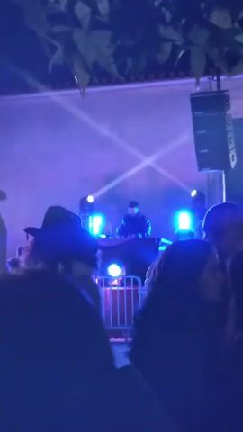 The video shows a DJ performing live on stage at an outdoor event. A crowd is visible in the foreground, silhouetted against the stage lights. The DJ, standing behind a set of equipment, is illuminated by blue and white lights. He is wearing a black jacket and appears to be speaking into a microphone. A large speaker array is positioned on the right side of the stage. Tree branches are visible at the top of the frame. The overall atmosphere appears energetic and festive.