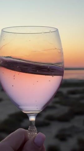 A close-up shot shows a wine glass being held in a hand with light pink nail polish. The glass contains a pink liquid that is sloshing. The background is blurred and shows a body of water and a beach with low-lying vegetation, all under a colorful sunset sky. The lighting and colors suggest it is either sunrise or sunset.