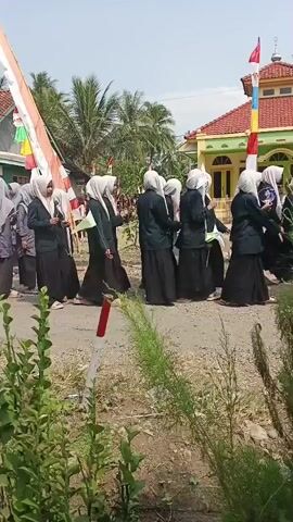 A group of women, all wearing hijabs and black skirts, walk down a dirt road. Most of them are wearing grey shirts, and one is wearing a black shirt. They are walking in a line, and some of them are carrying what appear to be long, thin objects. In the background, there are houses with red roofs and green accents, as well as palm trees. There are also flags and banners visible, suggesting some kind of celebration or event is taking place. The sky is clear and blue.