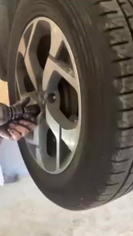 The video shows a person removing lug nuts from a car wheel with an impact wrench. The car is jacked up, the wheel is silver and black with a Kia logo on the center cap. The tire is black and appears worn. The person's hands are visible, and they are dirty. The environment is a garage or workshop, with a wooden wall in the background.