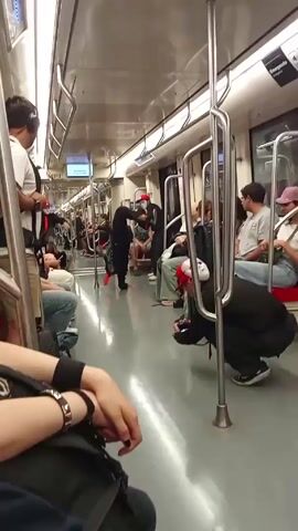 The video shows a group of street performers dancing in a metro car. They are dressed in black suits, red baseball caps, and white masks, and they perform breakdance moves to the music. Other passengers are seated on the side. The interior of the metro is bright and modern, with stainless steel handrails. A display screen shows the upcoming stop.