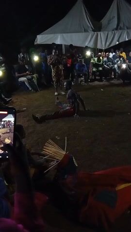 The video shows an outdoor performance at night. A person on the ground dressed in dark clothing and red pants is performing acrobatics. They roll around and appear to be doing push-ups with their legs in the air. Around the performer, people are seated on chairs, observing and filming. A person in the foreground is also filming with their phone, with a red and blue costume partially visible in the lower frame. Behind the audience are two large white tents. The scene is lit by spotlights.